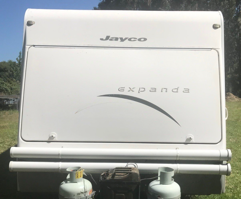 Rear view of a white Jayco Expanda motorhome or camper with two propane tanks attached at the bottom, photographed outdoors on grass with trees and blue sky in the background.