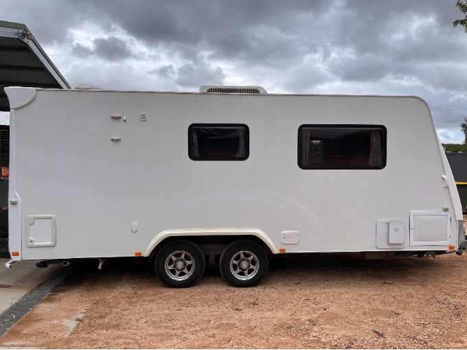 A white travel trailer with two windows and dual wheels, parked outdoors on a dirt ground under a cloudy sky.
