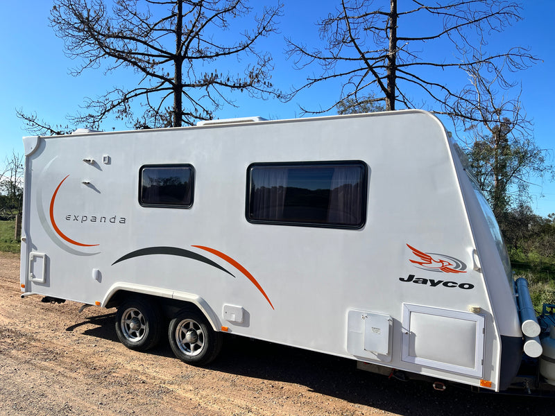 A white Jayco Expanda caravan parked outdoors on a dirt ground under a blue sky with bare trees. The side of the caravan features dark tinted windows, decorative orange and black swooshes, and the 'expanda' and 'Jayco' branding.