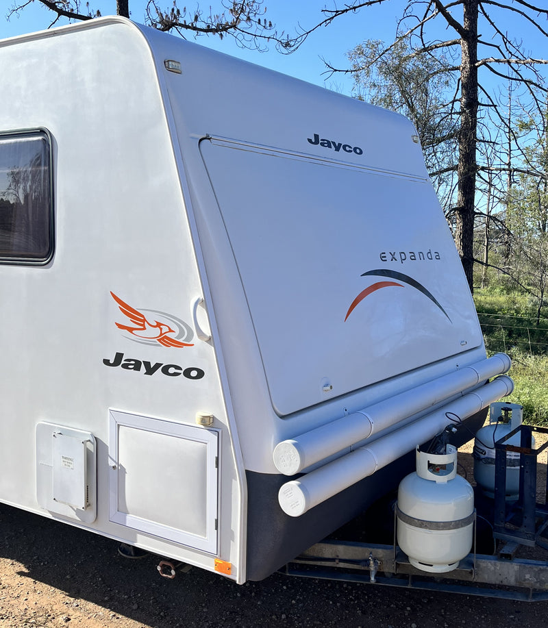 A white Jayco Expanda caravan parked outdoors on a gravel surface with trees and grass in the background. The side and rear of the caravan are visible, showing the Jayco branding, Expanda model name, storage compartments, and an external propane tank mounted below the rear.