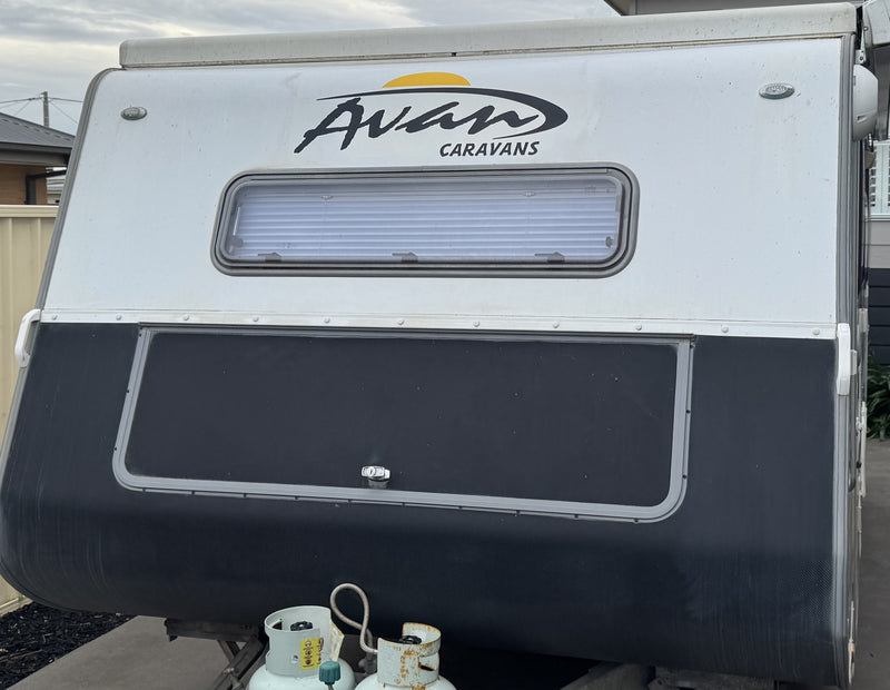 A front-facing view of a white and black Avan Caravans travel trailer, showing the front window, black front panel, and propane tanks connected at the bottom against an outdoor backdrop.