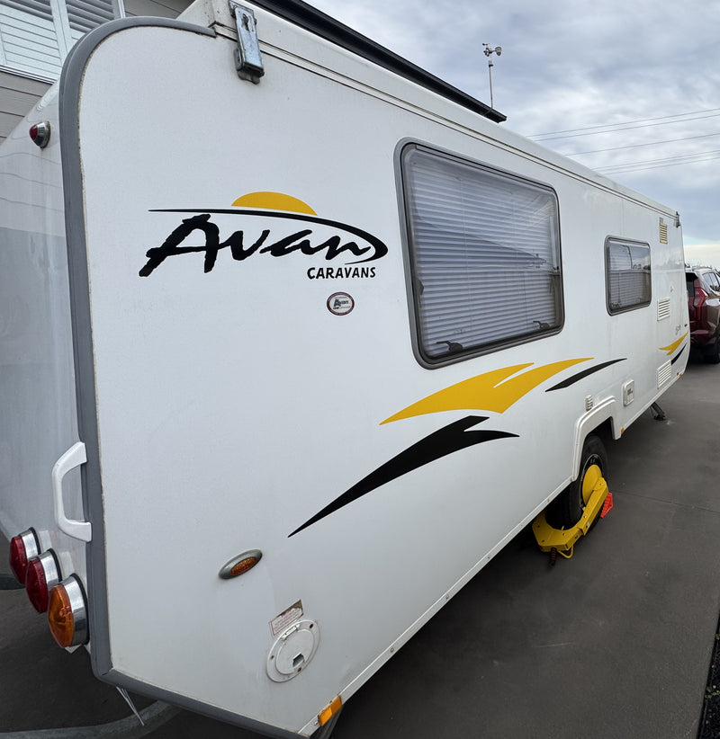 White Avan Caravans motorhome trailer parked outdoors on a paved surface, showing the rear and side exterior with windows, decals, and a yellow support leg extended.