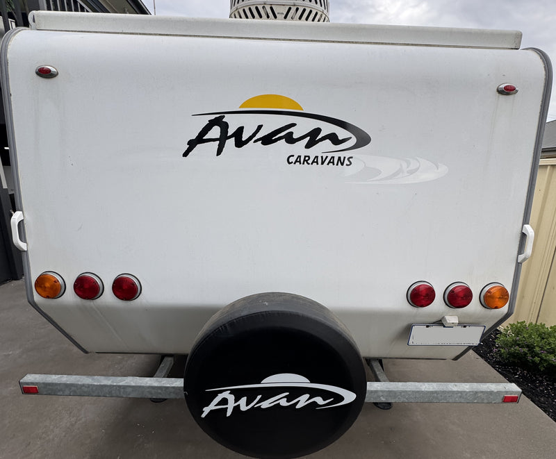 White Avan Caravans trailer rear view with a black spare tire cover, rear lights, and Avian logo decal on a concrete surface.