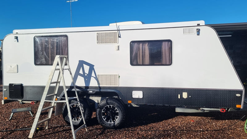 White caravan with a ladder and blue chair on a gravel surface under a clear blue sky.